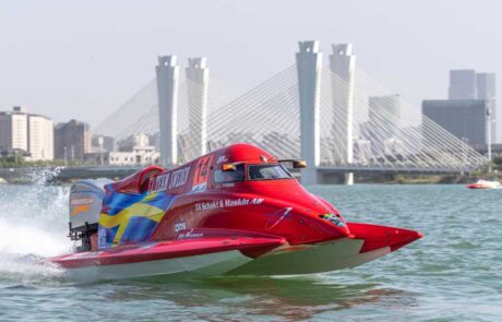 A red powerboat speeds through the water in front of a white suspension bridge and city in the distance.