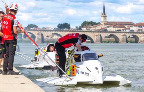 Race teams prepare two white powerboats lined up in the water in front of an old bridge in the distance.