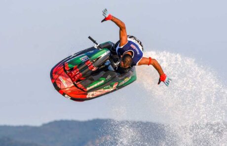 Racer spreads arms wide as his jet ski flys through the air leaving a trail of water spray behind it.