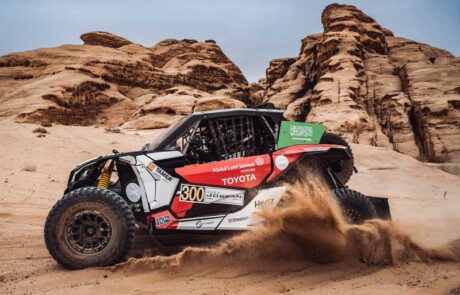 A Dakar rally car sprays up sand as it races past sand coloured rock formations in the desert.