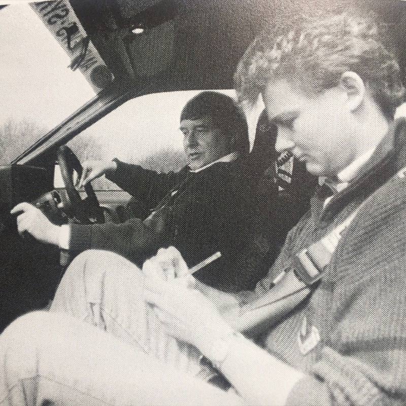 A black and white photo of Neil Perkins making notes whilst sat in the passenger seat next to a man talking at the steering wheel.