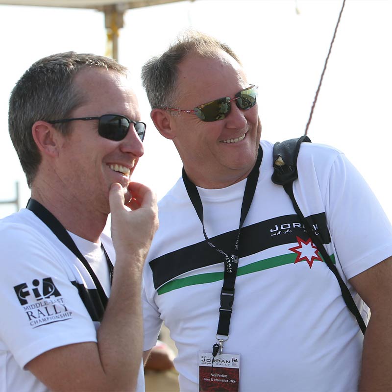 Neil Perkins and another man, both wearing white t-shirts and sunglasses, smile at something off camera.