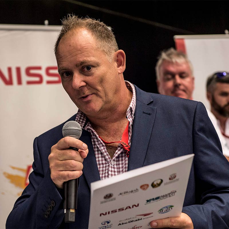 A photo of Neil Perkins wearing a suit and speaking into a microphone in front of a banner with the Nissan logo on it.